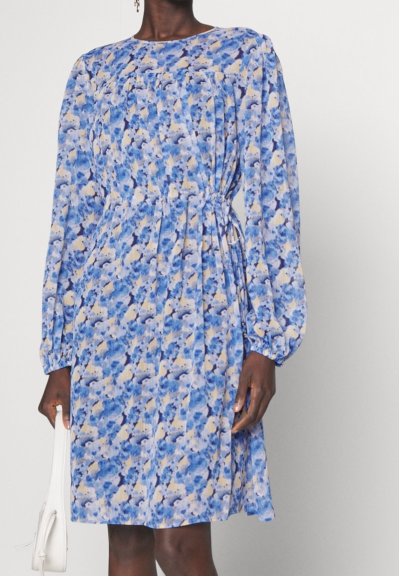 Femme portant une robe fleurie à manches longues et longueur genou dans des tons de bleu et beige, tenant un sac à main blanc à ses côtés.