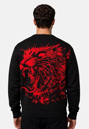 Hombre con suéter negro con un gran gráfico rojo de una cabeza de tigre rugiendo en la espalda, mostrado desde atrás sobre fondo liso.