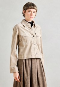 Veste beige raccourcie en tissu léger avec deux poches avant et fermeture à bouton. Associée à une jupe plissée marron.