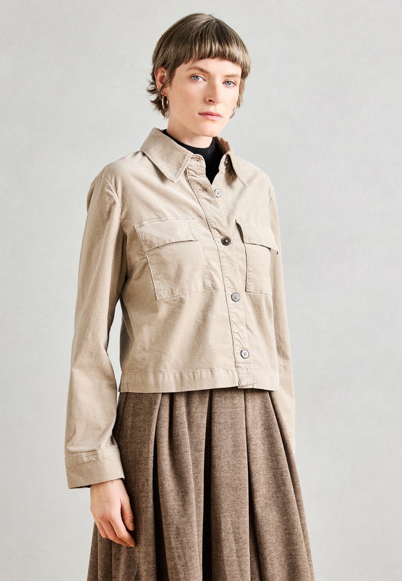 Veste beige raccourcie en tissu léger avec deux poches avant et fermeture à bouton. Associée à une jupe plissée marron.