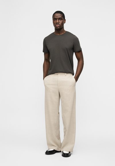 Άνδρας που στέκεται με τα χέρια στις τσέπες, φορώντας σκούρο γκρι μπλουζάκι, μπεζ παντελόνι με φαρδιά πόδια και μαύρα και λευκά παπούτσια σε απλό φόντο.