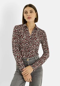 Blusa de manga larga en tela color crema con estampado de leopardo en rojo y negro. Cuenta con cuello y botones en la parte delantera. Combinada con jeans grises de cintura alta.