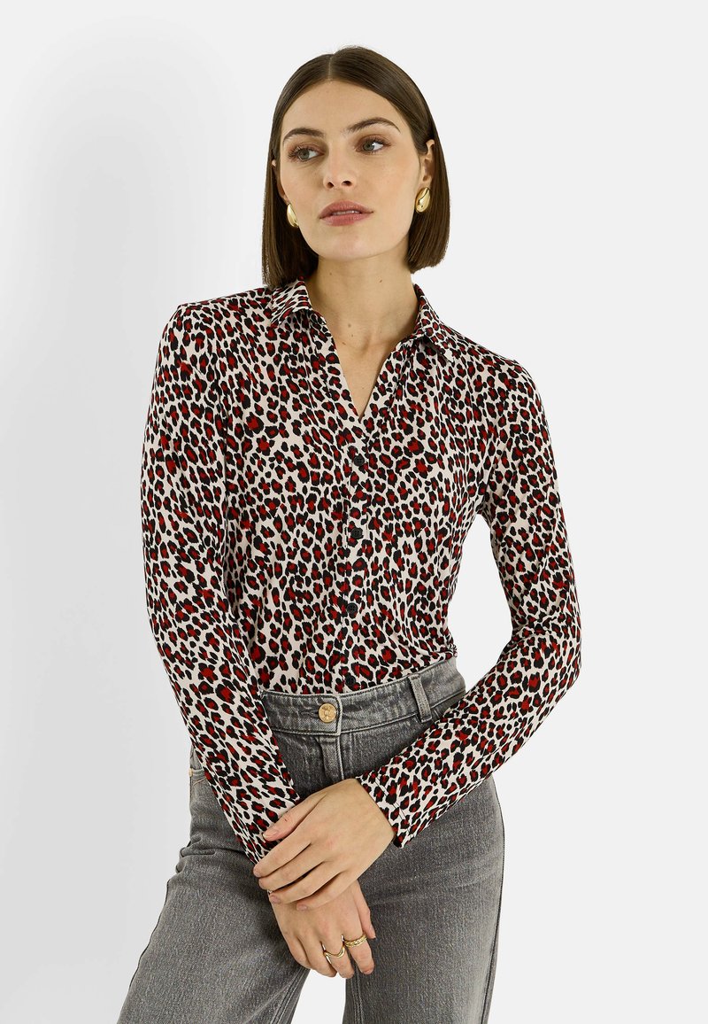 Blusa de manga larga en tela color crema con estampado de leopardo en rojo y negro. Cuenta con cuello y botones en la parte delantera. Combinada con jeans grises de cintura alta.