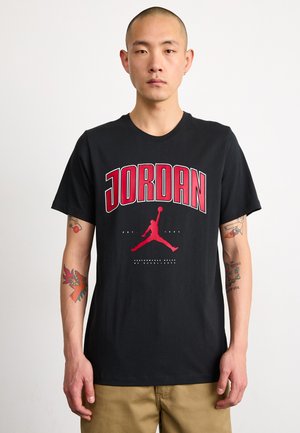 Sort bomulds t-shirt med fed rød "JORDAN" skrift, et ikonisk silhuetlogo, korte ærmer og rund hals.