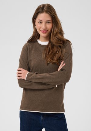 Jeune femme aux longs cheveux ondulés portant un pull marron par-dessus une chemise blanche, souriante, bras croisés devant un fond gris uni.