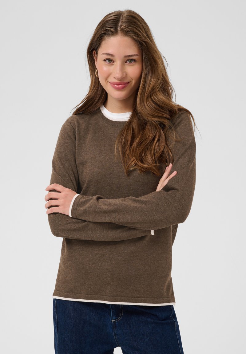 Jeune femme aux longs cheveux ondulés portant un pull marron par-dessus une chemise blanche, souriante, bras croisés devant un fond gris uni.