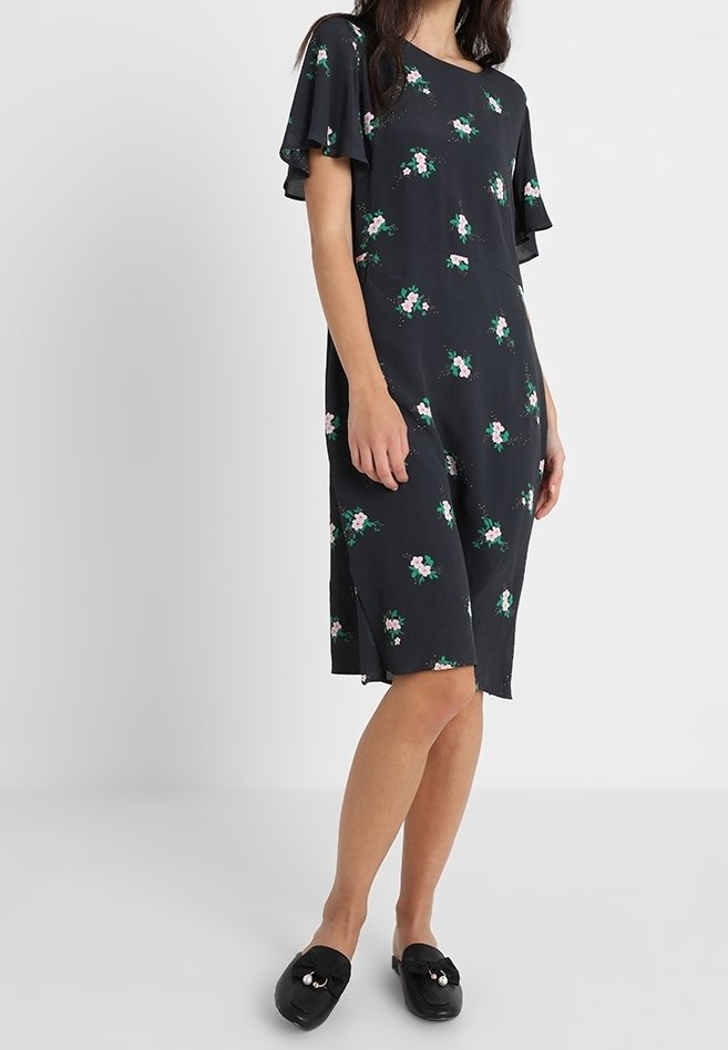 Femme portant une robe noire mi-longue avec un imprimé floral rose et vert et des manches volantées, associée à des chaussures noires slip-on avec des détails en perles.