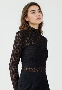 Maglietta nera a maniche lunghe, caratterizzata da un motivo in pizzo testurizzato con un colletto alto. Design aderente, che rivela un accenno di pelle attraverso il tessuto.