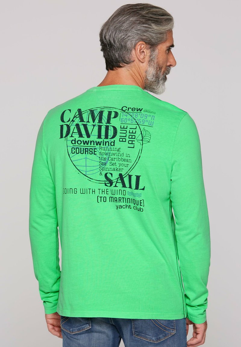 T-shirt a maniche lunghe verde con testo stampato e grafiche nere, che includono elementi nautici e lettering stilizzato. Tessuto in misto cotone.