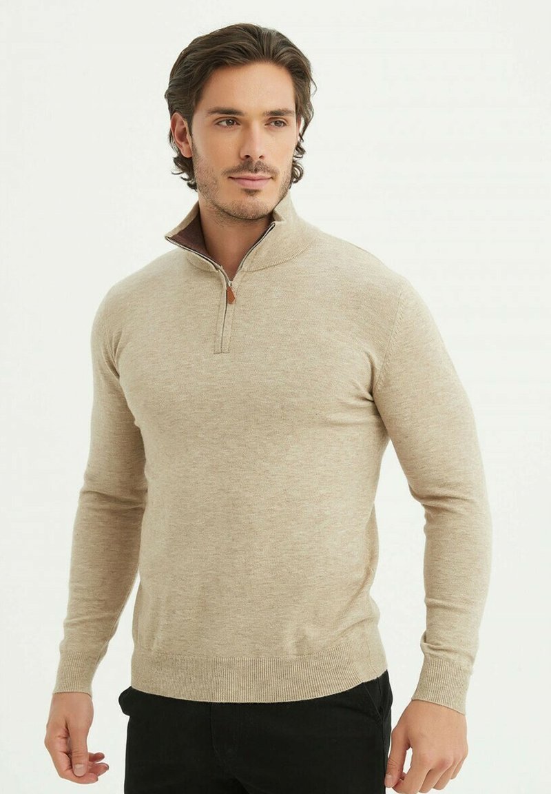 Pull en tricot beige avec un col zippé mi-long, présentant un ourlet et des poignets côtelés. Texture lisse, design ajusté et accent sombre sur le col.