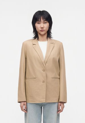Jeune personne aux cheveux noirs portant un blazer beige à deux boutons sur une chemise blanche et un jean bleu clair, debout devant un fond uni.