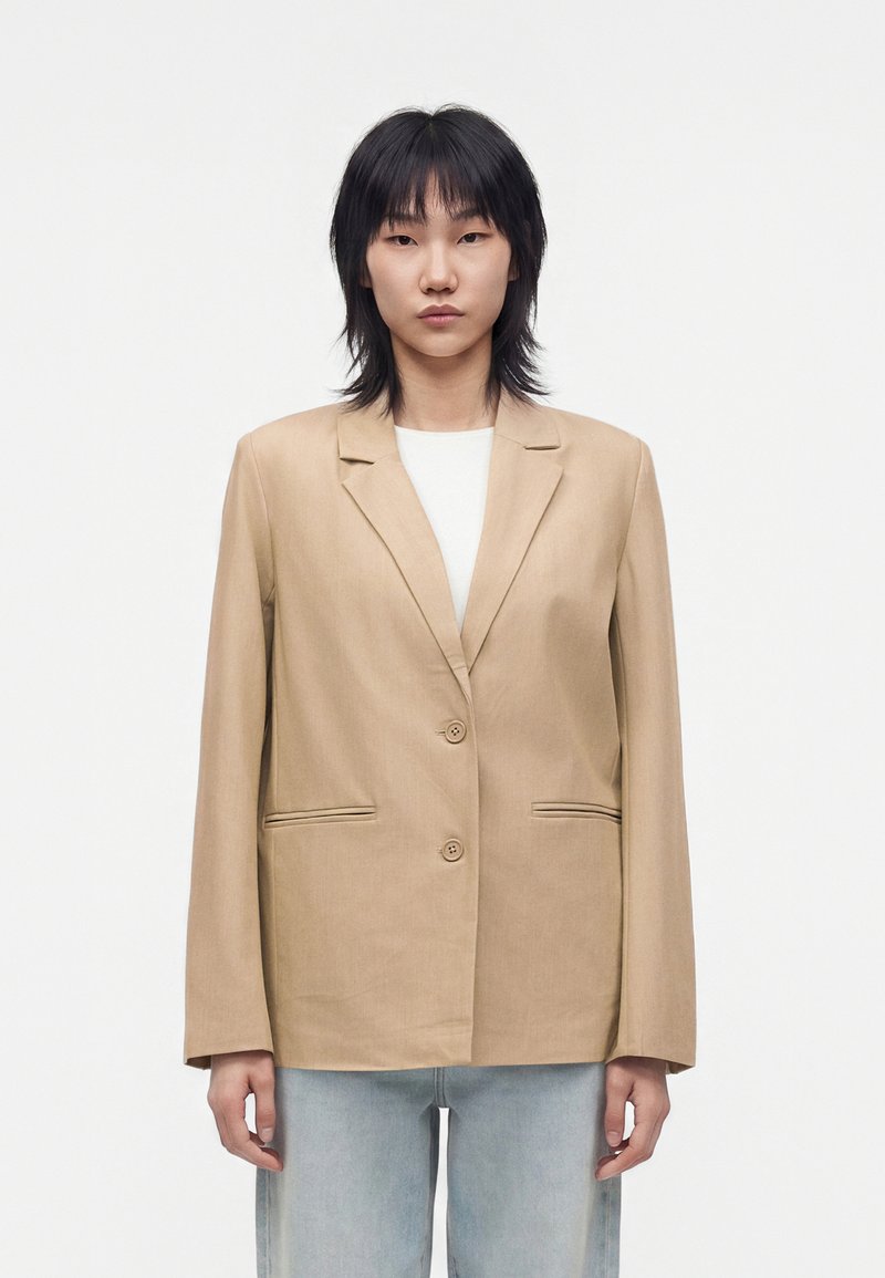 Jeune personne aux cheveux noirs portant un blazer beige à deux boutons sur une chemise blanche et un jean bleu clair, debout devant un fond uni.