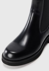 Stivale Chelsea in pelle nera lucida con punta arrotondata, pannello laterale elastico e suola in gomma nera su uno sfondo bianco.