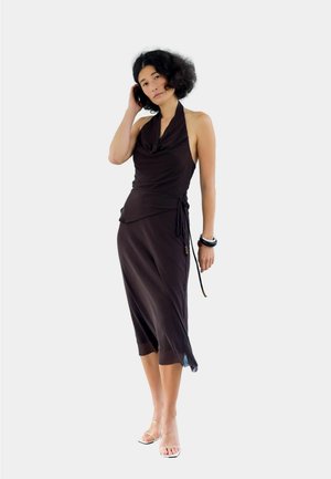 Femme aux cheveux bouclés portant une robe noire sans manches à col halter et des talons à brides transparentes, debout, une main touchant ses cheveux.