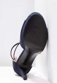 Chaussure à talons hauts en daim bleu marine inclinée vers le haut, montrant une semelle noire lisse et un talon fin sur fond d'angle blanc.