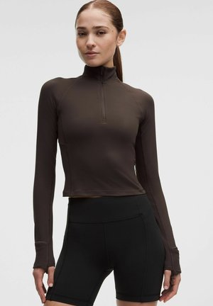 Vrouw draagt een aansluitend donkerbruin langarmig ritsvestje met duimgaten en zwarte high-waist shorts tegen een effen achtergrond.