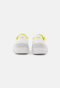 Witte sneakers met een gladde leren bovenkant, grijze suède accenten, felle gele veters en "HUGO" branding op de hiel. Plat rubberen zool.