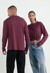 Langermede burgunder topper brukt med svarte og lyseblå jeans. Den kvinnelige toppen har en knyttet front; mannens topp er ensfarget.