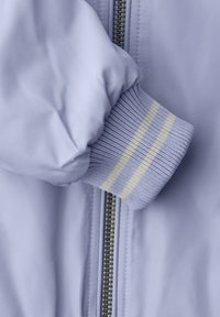 Manica di giacca viola chiaro con polsino a coste caratterizzato da due strisce crema, mostrata accanto a una cerniera argentata su tessuto abbinato.