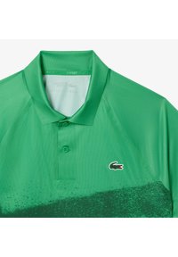 Grön pikétröja med en texturerad design, med ett gradiente mönster och en liten Lacoste-logotyp på vänster bröst. Tre-knapps knäppning.