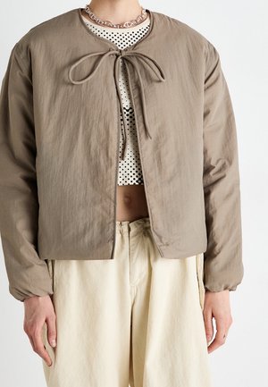 Persoon die een beige jas met strik aan de voorkant draagt over een wit cropped top met polkadots en losse crèmekleurige broek, met een zilveren ketting.
