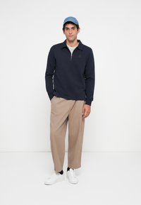 Man wearing blue cap, navy quarter-zip sweatshirt, beige wide-leg pants, black socks, and white sneakers against a plain white background.