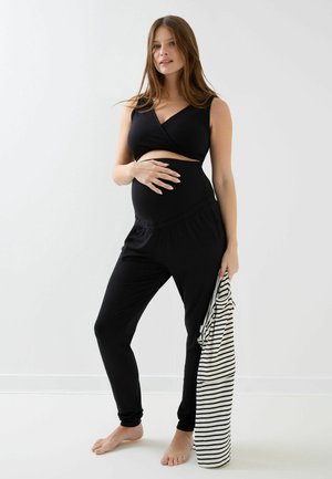 Kobieta w ciąży w czarnym stroju ciążowym trzyma paskowane ubranie, stojąc boso na jednolitym białym tle.