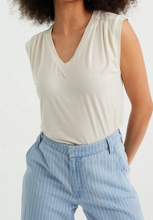 Kobieta w bezrękawniku w kolorze kości słoniowej z dekoltem w serek i jasnoniebieskich, wysokowiejskich spodniach w cienkie paski, z rękami w kieszeniach.