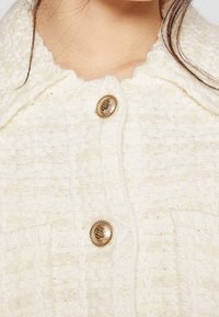 Gros plan d’un cardigan tricoté texturé crème avec un col festonné et deux boutons dorés ornés, porté par une personne avec des mèches de cheveux foncées.