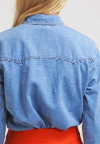 Ljusblå denimskjorta med krage, synliga stygn och avslappnad passform, kombinerad med ett texturerat rött midjeband.