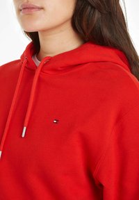 Kobieta w jasnoczerwonym hoodie z troczkami i małym prostokątnym logo na piersi, długie ciemne włosy widoczne na ramieniu.