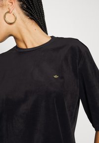 Svart T-shirt med kort ärm i mjukt tyg, med en guldbroderad logotyp på vänster bröst och en klassisk rund halsringning.