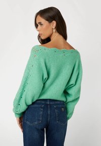 Maglione a maglia verde menta con una vestibilità ampia, scollatura a onde e dettagli a maglia aperta sulle maniche, abbinato a jeans blu scuro.