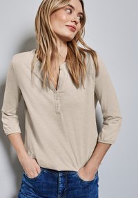 Beige Langarmoberteil mit Henley-Ausschnitt, das vier Knöpfe hat. Der Stoff ist weich, leicht strukturiert und wird mit blauen Jeans kombiniert.
