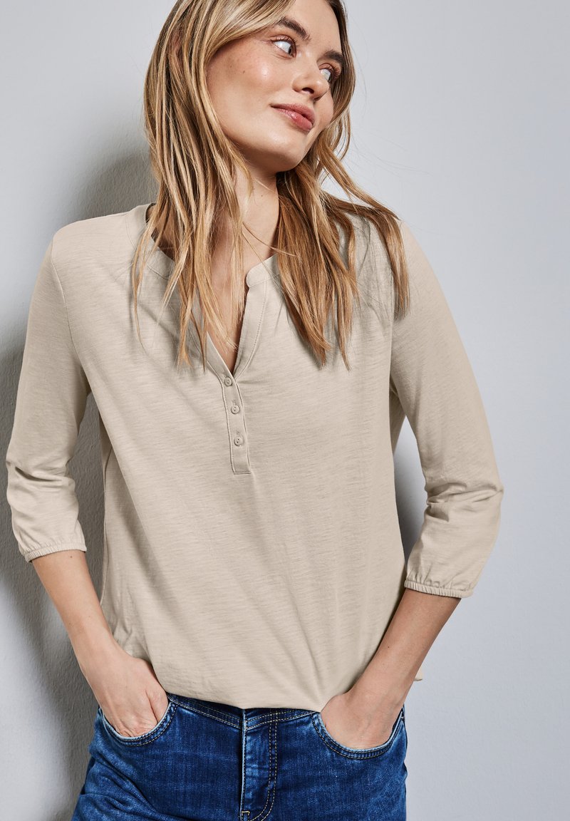 Beige Langarmoberteil mit Henley-Ausschnitt, das vier Knöpfe hat. Der Stoff ist weich, leicht strukturiert und wird mit blauen Jeans kombiniert.