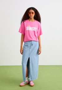 Růžové grafické tričko s nápisem "SUNSET", světle modrá denimová maxi sukně s předním rozparkem, růžové tenisky se zelenými pruhy a sluneční brýle.