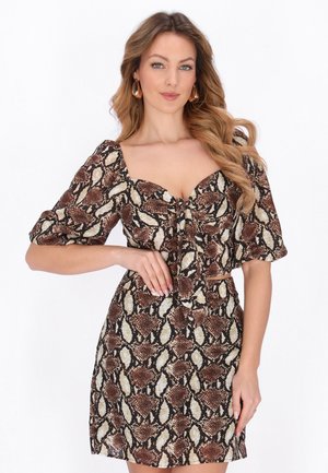 Kvinna som bär en klänning med brunt och beige ormmönster, puffärmar och knytning framtill, poserar med ena handen på höften.