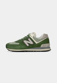 Žalios ir smėlinės New Balance sportbačiai su zomšos ir tinklelio medžiagomis, raišteliais per priekį ir paminkštinta ENCAP tarpu, šoninis vaizdas baltoje fone.