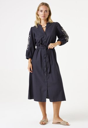 Robe boutonnée bleu marine foncé avec une taille ceinturée, des manches bouffantes et une broderie florale sur les manches. Fabriquée en tissu léger.