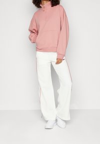 Sudadera con cremallera de color rosa claro, con un bolsillo tipo canguro, combinada con pantalones anchos blancos que tienen rayas rojas a los lados y zapatillas deportivas blancas.