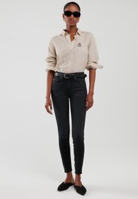 Žena u bež košulji s prugama na kopčanje, crnim uskim trapericama, crnim remenom, crnim ravnim cipelama sa špicom i crnim sunčanim naočalama, stoji nasuprot jednobojnoj pozadini.