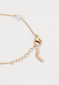 Gold-colored metal chain bracelet featuring a pearl bead, a lobster clasp, and a small engraved tag. Smooth texture with shiny finish.