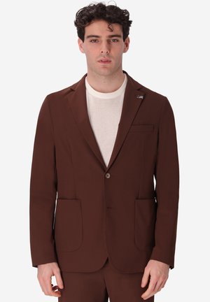 Uomo che indossa una giacca marrone sopra una maglietta bianca girocollo, in piedi contro uno sfondo bianco uniforme.