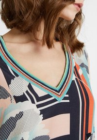 Multikleurige V-hals top met een patroonontwerp. Voorzien van een textuurafwerking in blauw, oranje en groen. Zachte stof met bloemen- en abstracte vormen.