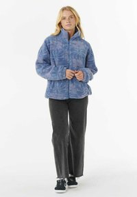 Veste en polaire bleue avec un motif texturé, col montant et fermeture éclair. Associée à un pantalon gris et des baskets noires.