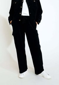 Schwarze Cordjacke mit Knopfverschluss und zwei Brusttaschen, kombiniert mit passenden Cargo-Hosen und weißen Sneakers. Glatte Textur.