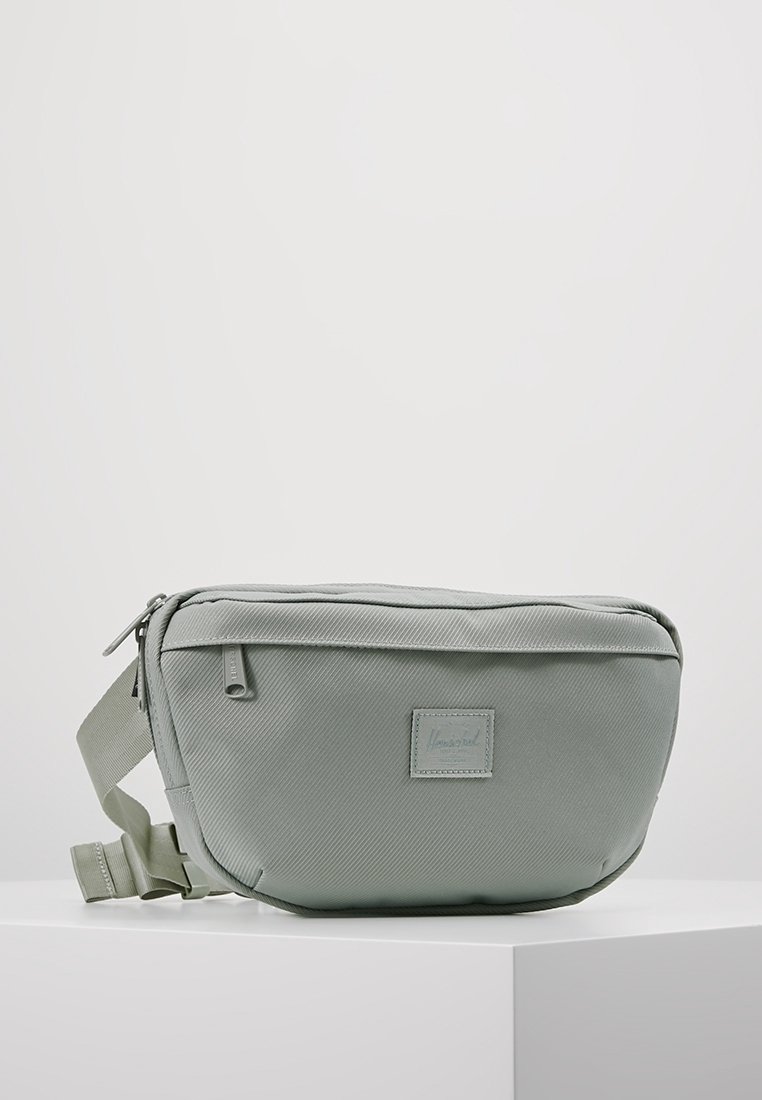 Sac bandoulière en tissu vert clair, présentant une texture lisse, une fermeture éclair supérieure, une sangle réglable et une petite étiquette de marque à l'avant.