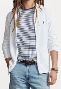 Vit zip-up hoodie som bärs över en blå- och vitrandig t-shirt. Matchas med ljusblå jeans och ett brunt flätat bälte, komplett med en klocka.