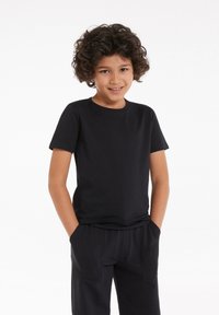 Usmívající se dítě s kudrnatými vlasy, oblečené v černém tričku s krátkým rukávem a černých kalhotách, stojící s rukama v kapsách na bílém pozadí.