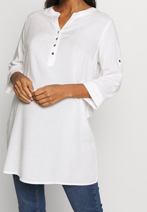 Femme portant une tunique blanche ample avec des manches trois-quarts, boutons noirs sur le devant et les manches, associée à un jean bleu.